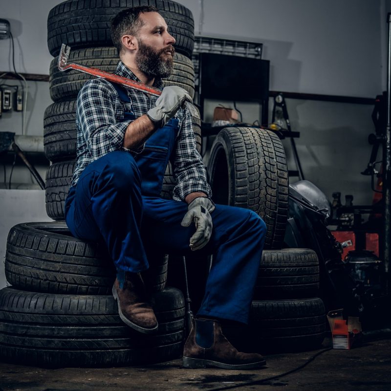 Bearded mechanic sits on an old car's tire in a garage.