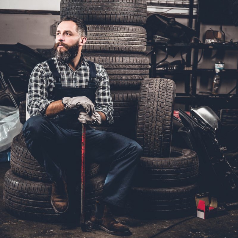 Portrait of bearded mechanical sits on an old tire in a garage.